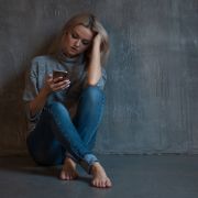 Blond woman sitting on floor in front of gray wall, sadly looking at phone