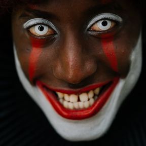 A clown with bright white contacts and red and white makeup smiles a sinister smile