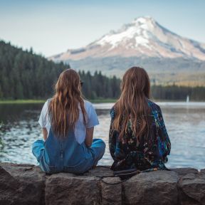Two women sitting on rock facing a lake and mountains.