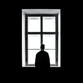 A black and white photo of a silhouette of a man staring out a window. 
