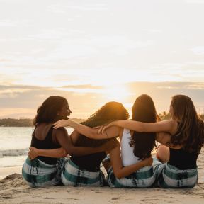 a group of four women sitting on the beach at sunset with arms around eachother 