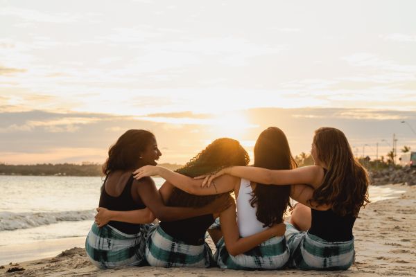 a group of four women sitting on the beach at sunset with arms around eachother 