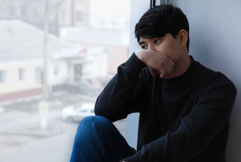 Sad young man looking out window, wearing dark clothes
